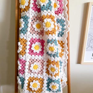 May include: A handmade crochet blanket featuring daisy flower motifs. The blanket is made with squares in white, pink, teal, and yellow, with white daisy appliqu&eacute;s. The blanket is draped over a wooden ladder.