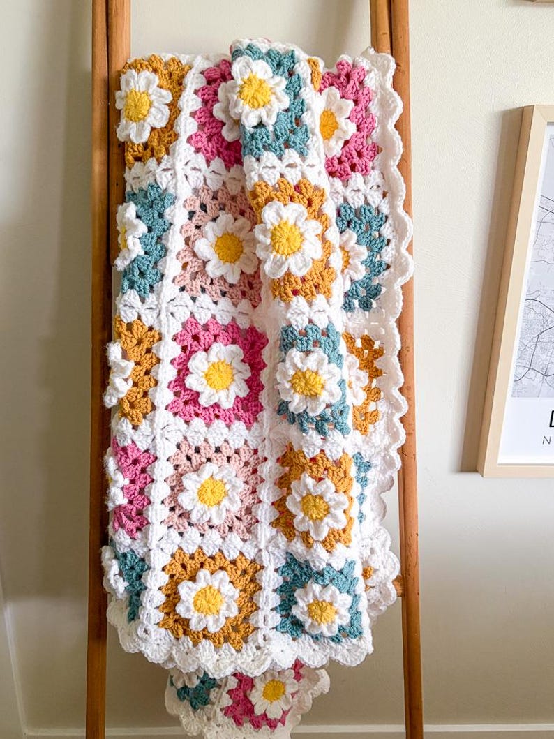 May include: A handmade crochet blanket featuring daisy flower motifs. The blanket is made with squares in white, pink, teal, and yellow, with white daisy appliqu&eacute;s. The blanket is draped over a wooden ladder.
