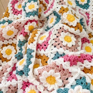 May include: A handmade crochet blanket featuring a daisy pattern. The blanket is made of white, pink, yellow, teal, and mustard-colored yarn. The daisy flowers are three-dimensional and add texture to the blanket.