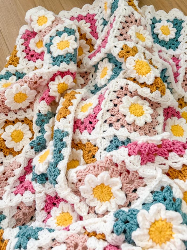 May include: A handmade crochet blanket featuring a daisy pattern. The blanket is made of white, pink, yellow, teal, and mustard-colored yarn. The daisy flowers are three-dimensional and add texture to the blanket.