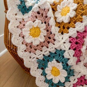 May include: A handmade crochet blanket with a daisy flower design. The blanket features white, yellow, pink, teal, and mustard yellow squares. The blanket is folded in a wicker basket. The blanket is made of soft yarn.