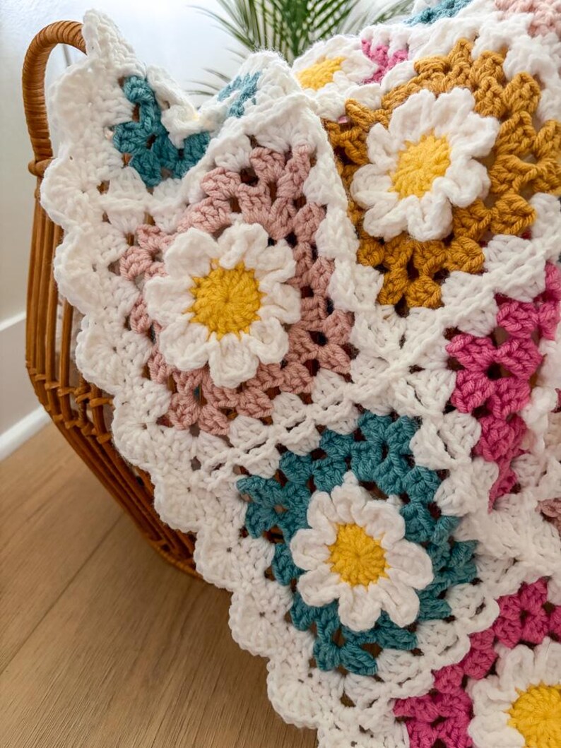 May include: A handmade crochet blanket with a daisy flower design. The blanket features white, yellow, pink, teal, and mustard yellow squares. The blanket is folded in a wicker basket. The blanket is made of soft yarn.