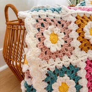 May include: A handmade crochet blanket with a daisy pattern. The blanket features squares in white, pink, teal, and yellow, with white daisy flowers in the center of each square. The blanket is draped over a wicker basket.