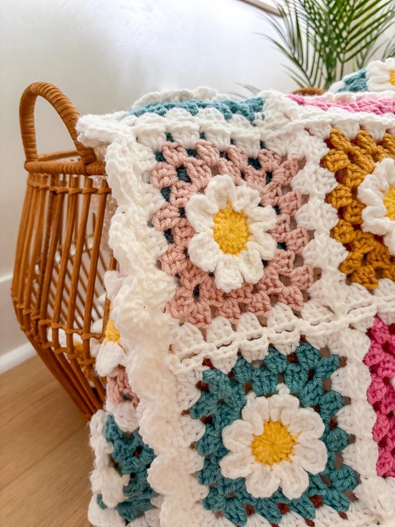 May include: A handmade crochet blanket with a daisy pattern. The blanket features squares in white, pink, teal, and yellow, with white daisy flowers in the center of each square. The blanket is draped over a wicker basket.