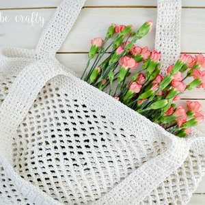 May include: A white crocheted mesh tote bag with a wide handle, filled with a bouquet of pink carnations. The bag is on a white wooden surface. "just be crafty" is written in white script on the top left corner of the image.