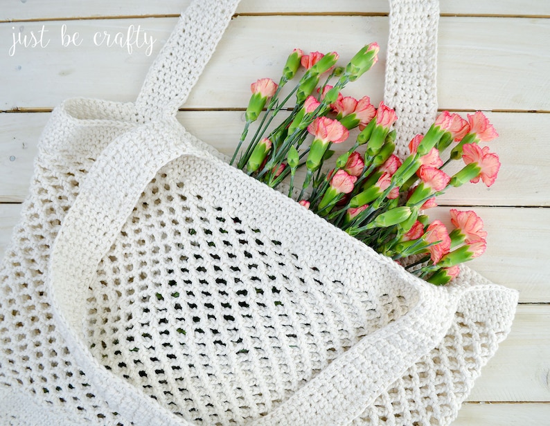 May include: A white crocheted mesh tote bag with a wide handle, filled with a bouquet of pink carnations. The bag is on a white wooden surface. "just be crafty" is written in white script on the top left corner of the image.