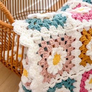 May include: A handmade crochet blanket with a daisy pattern in white, pink, yellow, and teal. The blanket is folded in a wicker basket, showcasing the intricate stitchwork and vibrant colors. The blanket is a cozy home decor item.