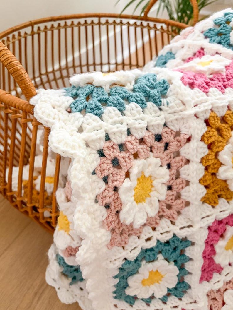 May include: A handmade crochet blanket with a daisy pattern in white, pink, yellow, and teal. The blanket is folded in a wicker basket, showcasing the intricate stitchwork and vibrant colors. The blanket is a cozy home decor item.