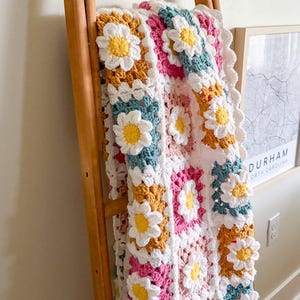 May include: A colorful, handmade crochet blanket featuring a daisy pattern. The blanket is made with white, yellow, pink, teal, and mustard-colored yarn. It is draped over a wooden ladder, showcasing its intricate design.