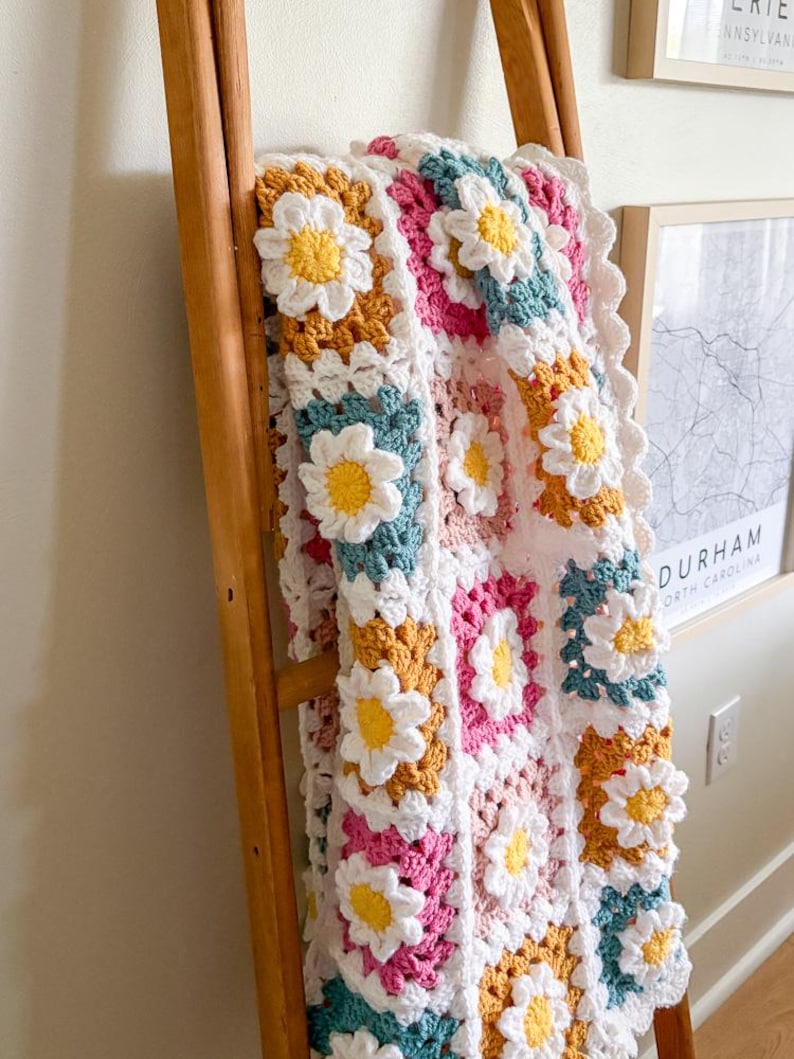 May include: A colorful, handmade crochet blanket featuring a daisy pattern. The blanket is made with white, yellow, pink, teal, and mustard-colored yarn. It is draped over a wooden ladder, showcasing its intricate design.