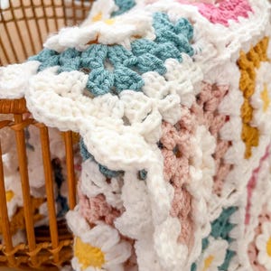 May include: A handmade crochet blanket with a daisy pattern. The blanket features white, light blue, pink, and yellow flowers. The blanket is folded over a wicker basket, showcasing the intricate stitchwork and color palette.