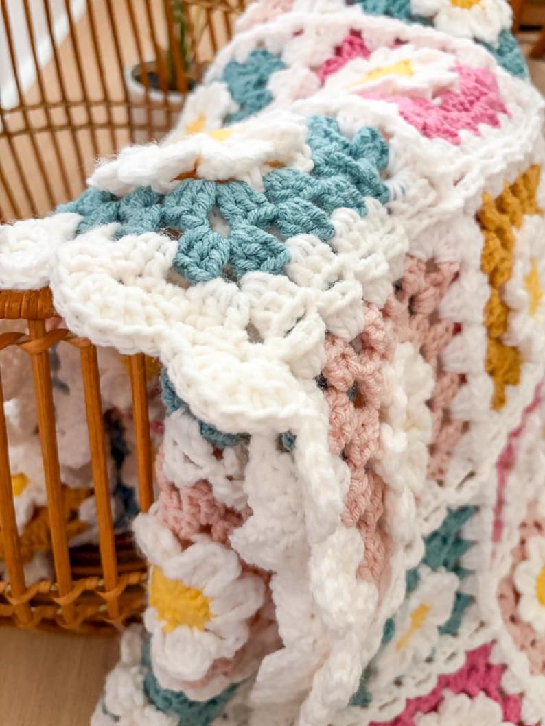 May include: A handmade crochet blanket with a daisy pattern. The blanket features white, light blue, pink, and yellow flowers. The blanket is folded over a wicker basket, showcasing the intricate stitchwork and color palette.