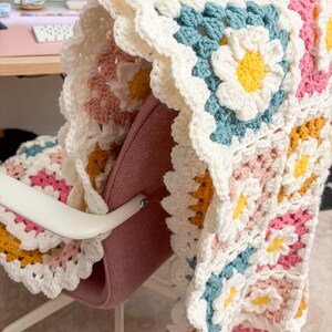 May include: A handmade crochet blanket draped over a chair. The blanket features a daisy pattern with white, yellow, pink, and blue flowers. The edges are scalloped, and the blanket is made of soft yarn.