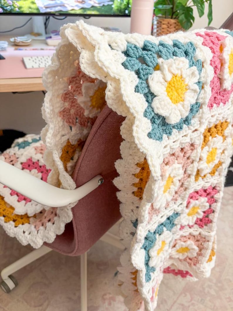 May include: A handmade crochet blanket draped over a chair. The blanket features a daisy pattern with white, yellow, pink, and blue flowers. The edges are scalloped, and the blanket is made of soft yarn.