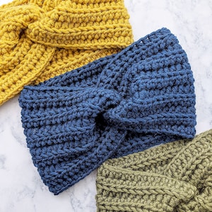 May include: Three crocheted headbands in different colors: mustard yellow, navy blue, and olive green. The headbands are all twisted and knotted in the center.