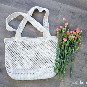 May include: A white crocheted tote bag with a wide, open top and a criss-cross strap design. The bag is sitting on a wood floor with a bouquet of pink carnations.