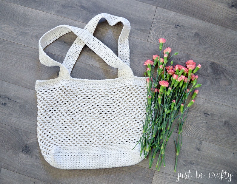 May include: A white crocheted tote bag with a wide, open top and a criss-cross strap design. The bag is sitting on a wood floor with a bouquet of pink carnations.