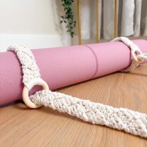 May include: A rolled-up pink yoga mat with a cream macrame strap and wooden ring closures. The strap is on a wooden floor. The yoga mat is a solid colour. The macrame strap is made of woven cotton.