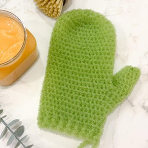 May include: A bright green, textured mitt with a loop, likely for use in the kitchen or bathroom. The mitt is surrounded by a wooden handled brush and a jar of orange-colored product, suggesting a cleaning or self-care theme.
