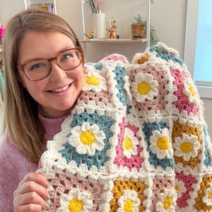 May include: A handmade crochet blanket featuring a daisy pattern. The blanket is made with white, yellow, pink, blue, and gold yarn. The daisy flowers are in the center of each square. The blanket is a cozy and colorful home decor item.
