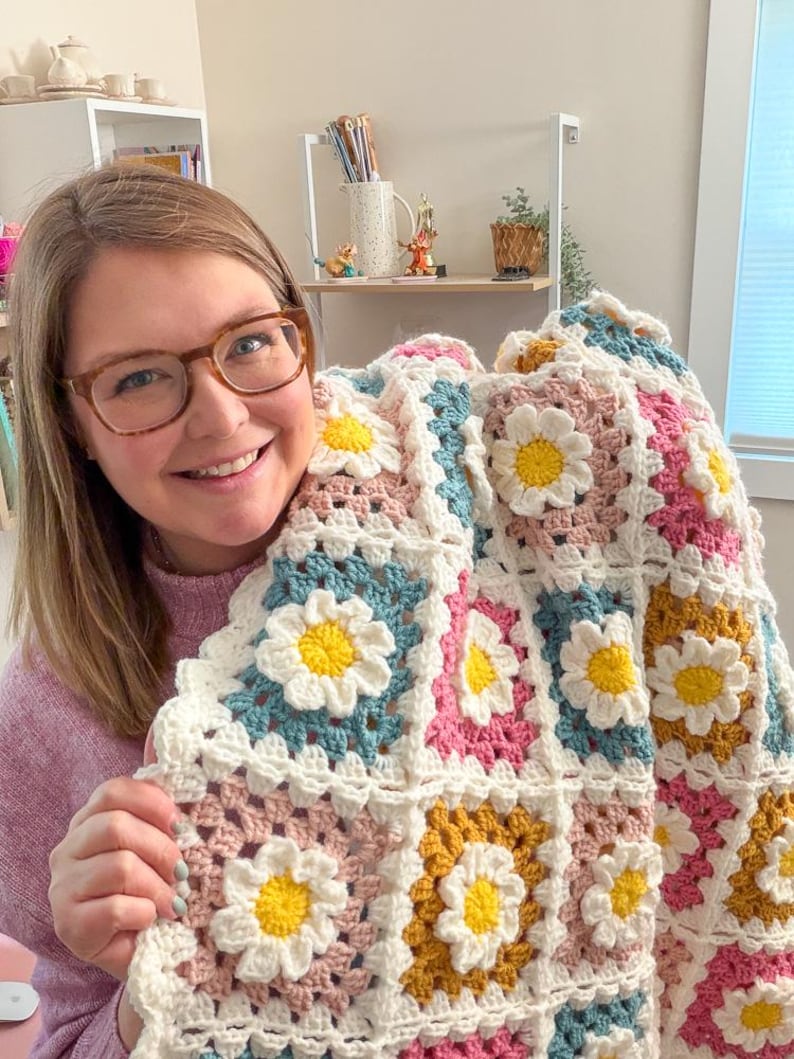 May include: A handmade crochet blanket featuring a daisy pattern. The blanket is made with white, yellow, pink, blue, and gold yarn. The daisy flowers are in the center of each square. The blanket is a cozy and colorful home decor item.