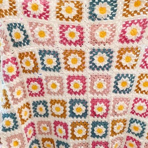 May include: A handmade crochet blanket featuring a daisy pattern. The blanket is composed of square blocks in white, pink, blue, and gold, each with a yellow daisy center. The edges are finished with a scalloped border.