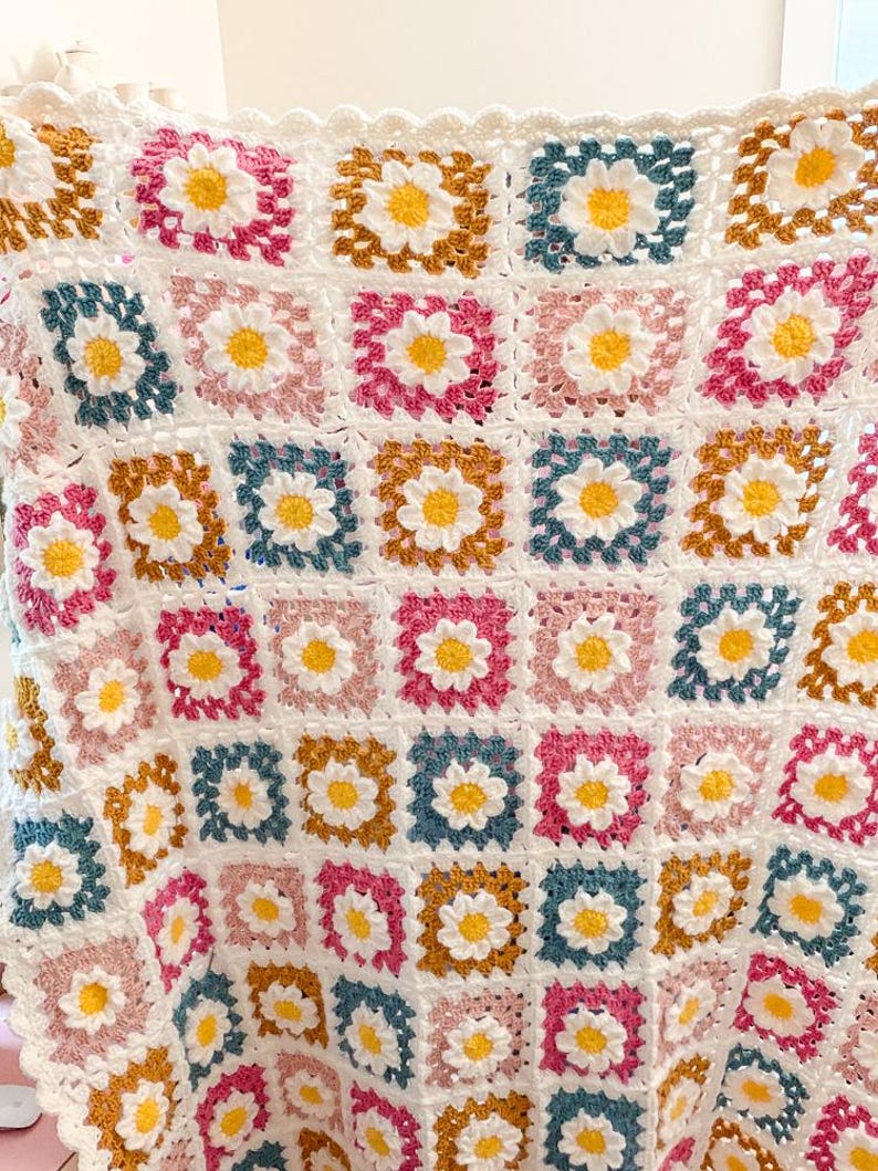 May include: A handmade crochet blanket featuring a daisy pattern. The blanket is composed of square blocks in white, pink, blue, and gold, each with a yellow daisy center. The edges are finished with a scalloped border.