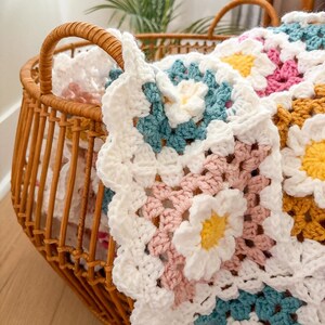 May include: A handmade crochet blanket with a daisy pattern in white, pink, yellow, and teal. The blanket is folded in a wicker basket with handles. The basket is made of woven brown material and sits on a wooden floor.