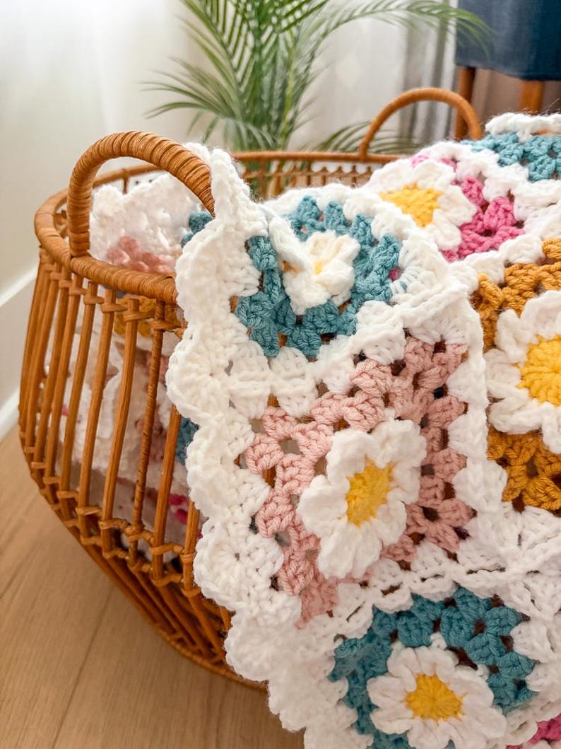 May include: A handmade crochet blanket with a daisy pattern in white, pink, yellow, and teal. The blanket is folded in a wicker basket with handles. The basket is made of woven brown material and sits on a wooden floor.