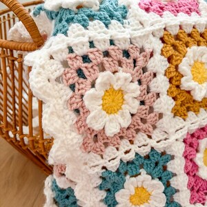 May include: A handmade crochet blanket with a daisy flower pattern. The blanket features white, pink, teal, and yellow squares. The blanket is folded in a wicker basket.