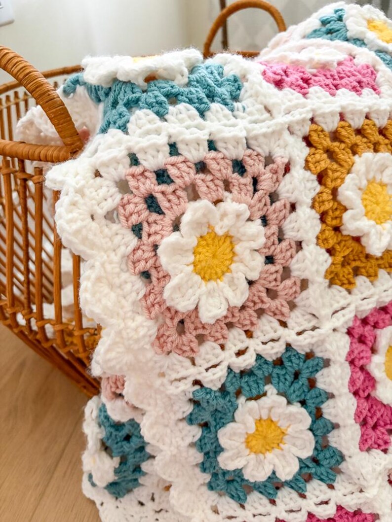 May include: A handmade crochet blanket with a daisy flower pattern. The blanket features white, pink, teal, and yellow squares. The blanket is folded in a wicker basket.