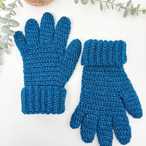 May include: A pair of handmade, blue crochet gloves with a textured appearance. The gloves have a ribbed cuff and a sparkly thread woven into the yarn. The gloves are displayed on a white surface with decorative greenery.