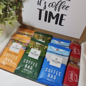 May include: A brown cardboard box with a white card on top that says "Rise & Shine it's coffee time". The box is open and contains several coffee bags and two packages of Lotus Biscoff cookies. The coffee bags are all from Taylors of Harrogate and are labeled with different roasts and flavors, including "Flying Start", "Rich Italian", and "Decaffe".