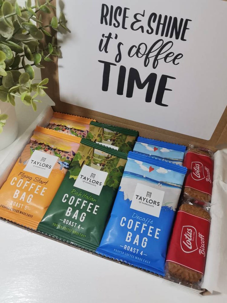 May include: A brown cardboard box with a white card on top that says "Rise & Shine it's coffee time". The box is open and contains several coffee bags and two packages of Lotus Biscoff cookies. The coffee bags are all from Taylors of Harrogate and are labeled with different roasts and flavors, including "Flying Start", "Rich Italian", and "Decaffe".