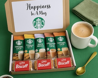 May include: A gift box with Starbucks coffee packets and Biscoff cookies. The box contains Caramel, Vanilla, White Mocha, Cinnamon Dolce, and Caramel Latte packets. A white mug of coffee and a gold spoon are on a green surface.