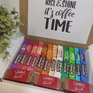 May include: A white gift box with a brown lid. The lid has a white card with the text "Rise & Shine it's coffee TIME". The box is filled with a variety of individually wrapped coffee flavored candy bars and two Lotus Biscoff cookies.