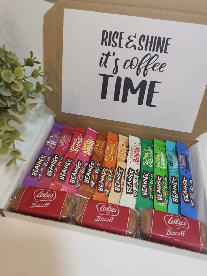 May include: A white gift box with a brown lid. The lid has a white card with the text "Rise & Shine it's coffee TIME". The box is filled with a variety of individually wrapped coffee flavored candy bars and two Lotus Biscoff cookies.