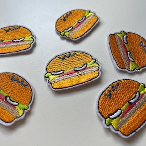 Pode incluir: Seis patches bordados termocolantes de hambúrgueres amarelos e castanhos com alface verde, carne rosa e sementes de sésamo brancas.