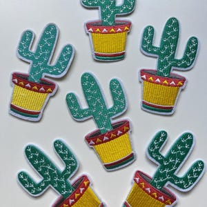Pode incluir: Seis cactos verdes em vasos amarelos com detalhes vermelhos e brancos. Os cactos são bordados em tecido branco.