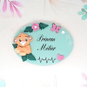 Könnte beinhalten: Ein hellblaues, ovales Namensschild mit einem Löwenjungen, Blumen, einem Schmetterling und einem Herzen. Das Schild trägt die Aufschrift "Prénom Métier" in schwarzer Kursivschrift.