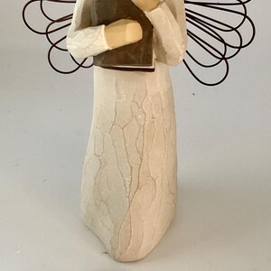 May include: A cream-colored angel figurine holding a brown book. The angel has a light brown head and wire wings. The figurine is standing on a white surface. The tag at the bottom reads "Angel of Learning."