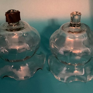 May include: Two clear glass candle holders with a bulbous shape and scalloped bases. One has a dark brown stopper. The candle holders are on a light blue surface. The glass has a vintage aesthetic.