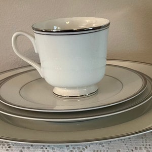 May include: A set of three white ceramic plates with a thin silver rim. The plates are stacked on top of each other, with a white ceramic teacup with a silver rim on top of the stack.