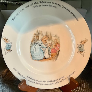 May include: White ceramic plate featuring a scene from a children's story. The plate has illustrations of rabbits and text around the rim. The central image shows a rabbit in a blue dress with other rabbits. The text reads: 'Now my dears,' said old Mrs. Rabbit...