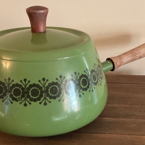 May include: Green enamel fondue pot with a wooden handle and lid. The pot has a black floral design around the rim.