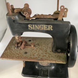 May include: Vintage Singer sewing machine in black with gold lettering. The machine has a rusty, aged appearance, with a metal base and a hand wheel on the side. The word "SINGER" is prominently displayed on the machine.