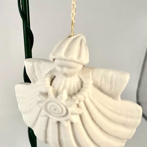 May include: A white ceramic angel Christmas ornament. The angel has a hat, wings, and a floral design on the front. The ornament is hanging from a gold string. The angel is approximately 4 inches tall.