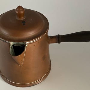 May include: A copper coffee pot with a wooden handle and a lid. The pot has a spout on the side and a small handle on the lid.