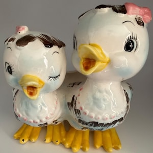May include: A pair of ceramic duck figurines with blue and white painted details. The ducks have yellow beaks and feet, brown eyes, and pink bows.