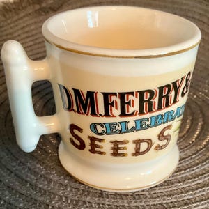 May include: Cream-colored ceramic mug with a curved handle and a gold rim. The mug features the text "DM FERRY & CELEBRATED SEEDS" in a vintage font. The mug has a slightly flared base and a smooth, glossy finish.